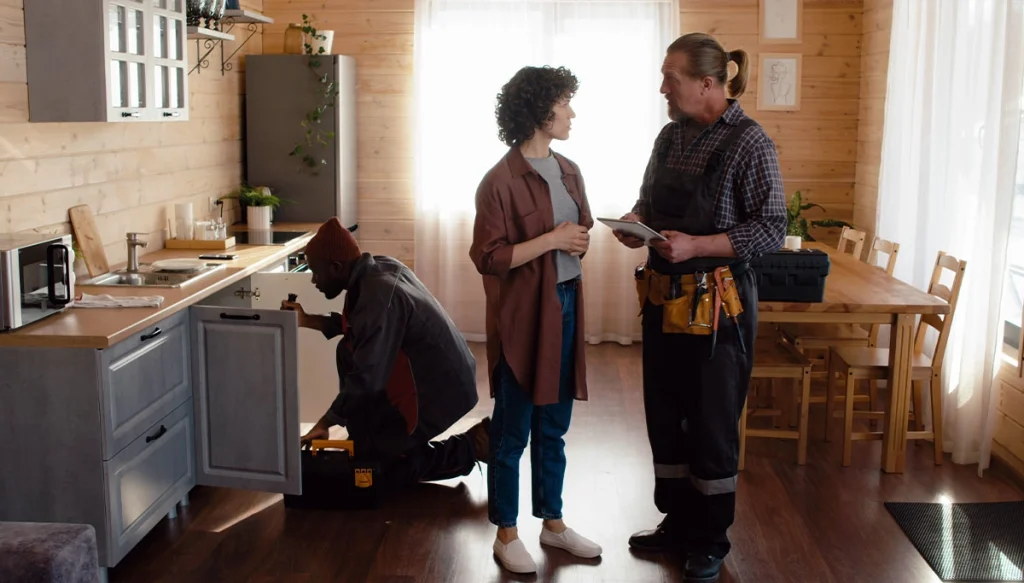 woman talking to a plumber while a second plumber checks the pipe under the sink