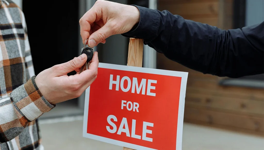 Two hands exchange keys underneath a home for sale sign